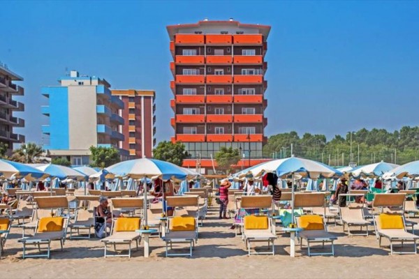 Písečná pláž s lehátky a slunečníky před moderním hotelem.