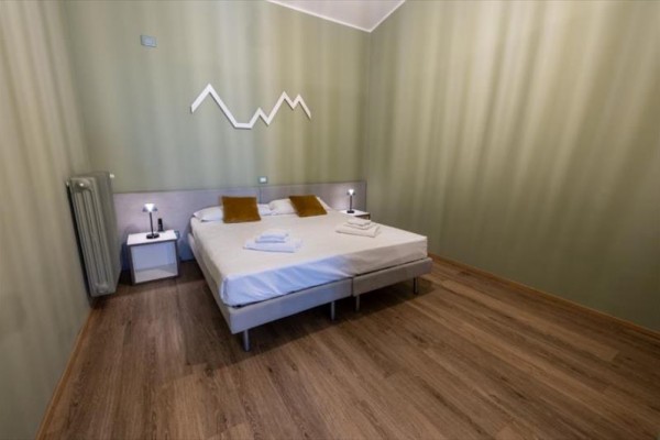 Ložnice s dvojlůžkem, bílými nočními stolky a dekorací ve tvaru hor na zdi.