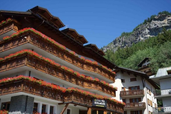 Dřevěný horský hotel s květinami na balkonech, v pozadí les a skála.
