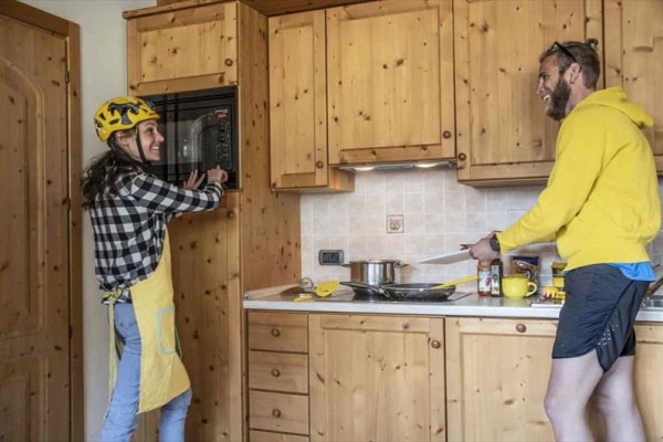 Dva lidé v kuchyni, jedna osoba s cyklistickou helmou a druhá s vařečkou.