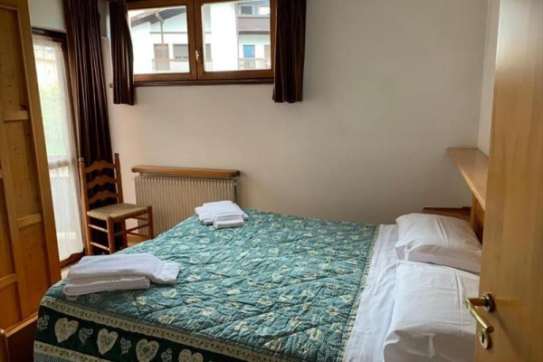 Ložnice s manželskou postelí pokrytou vzorovanou přikrývkou, dvěma složenými ručníky a dřevěnou židlí u okna.