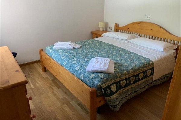 Ložnice s dřevěnou manželskou postelí, modrozeleným přehozem a složenými ručníky.