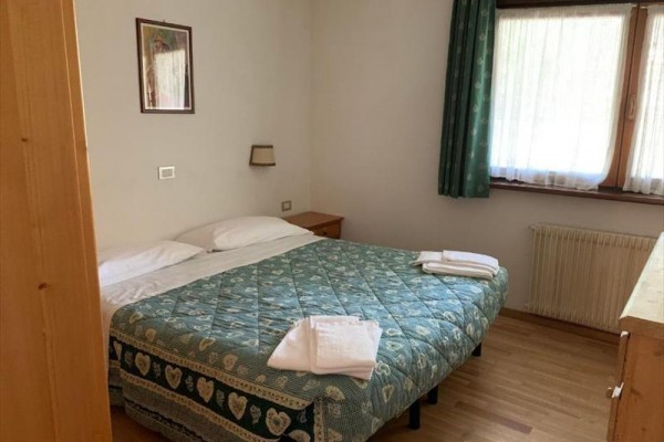 Ložnice s dvojlůžkem, zeleným přehozem, nočním stolem a oknem se závěsem.