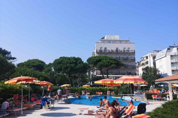 Lidé relaxují u bazénu s lehátky a slunečníky, v pozadí jsou stromy a hotel.