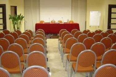 Konferenční místnost s řadami prázdných židlí a stolem s mikrofony.