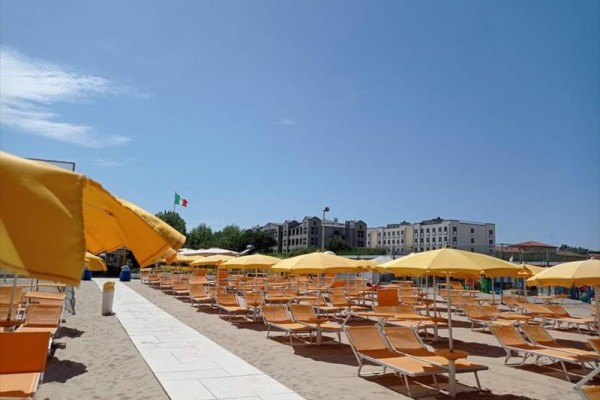 Pláž s řadami oranžových lehátek a žlutými slunečníky, v pozadí budovy a italská vlajka.