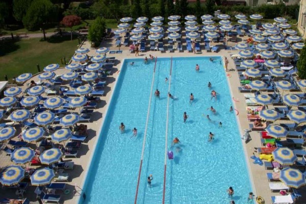 Venkovní bazén s lidmi obklopený řadami lehátek a slunečníků.  
