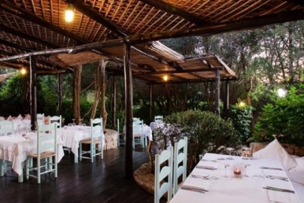 Venkovní restaurace s prostřenými stoly pod dřevěnou pergolou obklopenou zelení.
