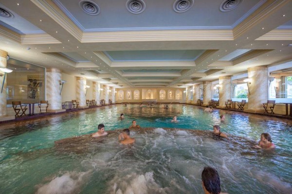 Lidé relaxující v krytém bazénu s vířivkou a sloupy po stranách.