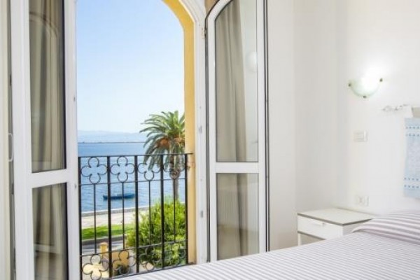 Ložnice s otevřenými dveřmi na balkon a výhledem na moře a palmu.
