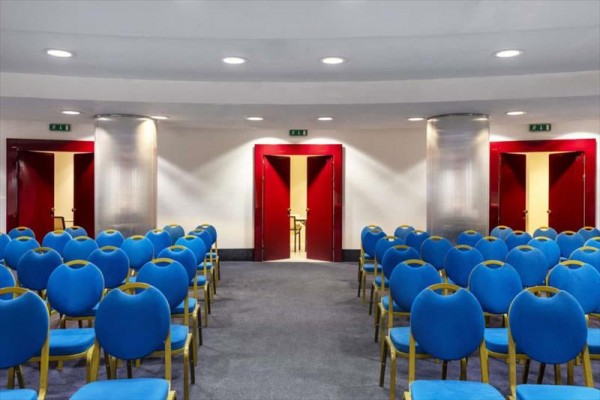 Konferenční místnost s modrými židlemi a červenými dveřmi vzadu.  