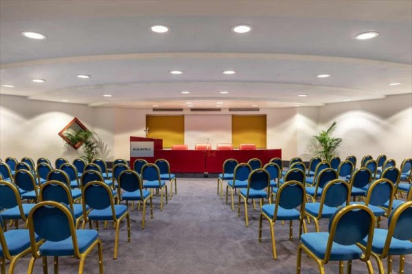 Konferenční místnost s řadami modrých židlí a červeným stolem v čele.