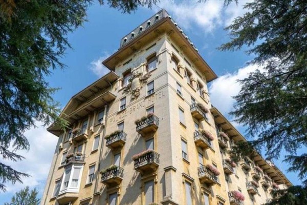 Historická budova hotelu s balkony a ozdobnými detaily pod modrou oblohou mezi stromy.  