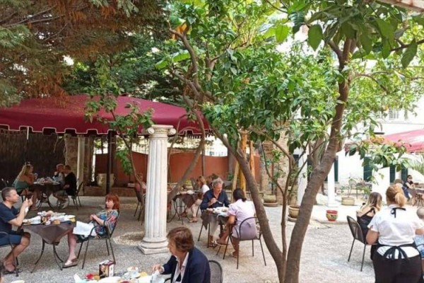 Lidé sedí a jedí v zahradní restauraci pod stromy a červenými slunečníky.