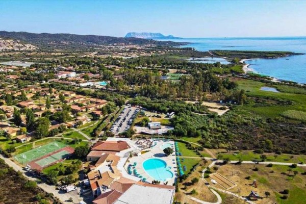 Letecký pohled na pobřežní oblast s resortem, bazénem a sportovištěm obklopené zelení.