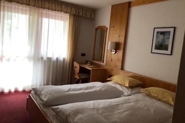 Dvojlůžkový pokoj s bílými povlečením, dřevěným nábytkem a oknem se záclonami.  