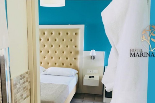 Pokoj s modrou stěnou, postelí s polstrovaným čelem a logem hotelu Marina del Capo.