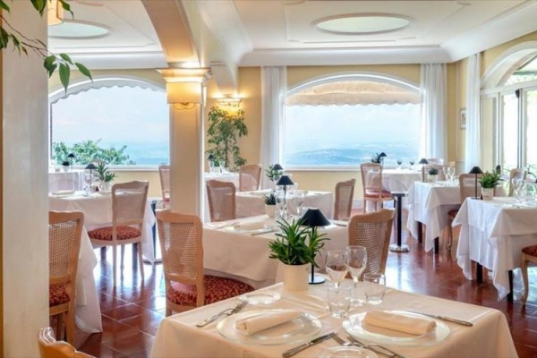Elegantní restaurace s bílými ubrusy a výhledem na moře skrz velká okna.