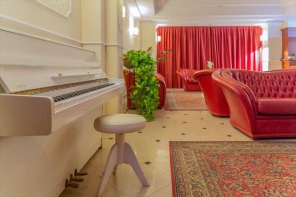 Bílé piano a červená kožená křesla v elegantním pokoji s červenými závěsy.