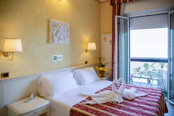 Elegantní hotelový pokoj s manželskou postelí a ručníky ve tvaru labutí na přikrývce, balkon s výhledem na moře.