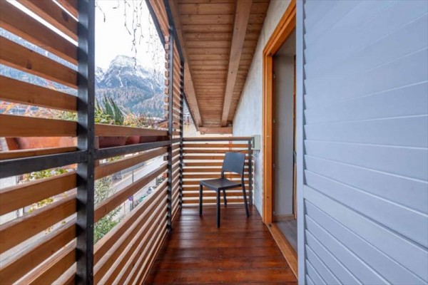 Dřevěný balkon s černou židlí a výhledem na zasněžené hory.