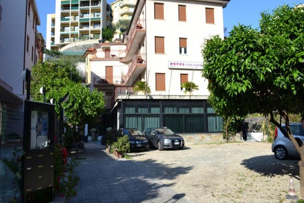 Parkoviště vedle hotelu s několika zaparkovanými auty a stromem.