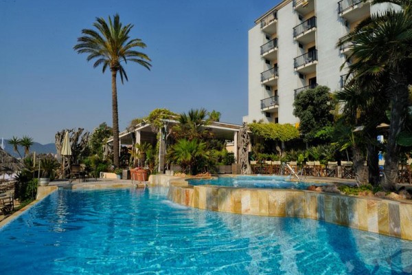 Hotel s bazénem obklopený palmami a rostlinami pod modrou oblohou.