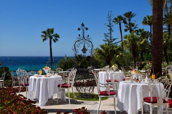 Venkovní restaurace s bílými stoly a výhledem na moře, obklopená palmami.  