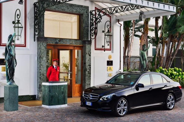 Vrátný v červené uniformě stojí u vchodu do budovy vedle černého vozu Mercedes-Benz.