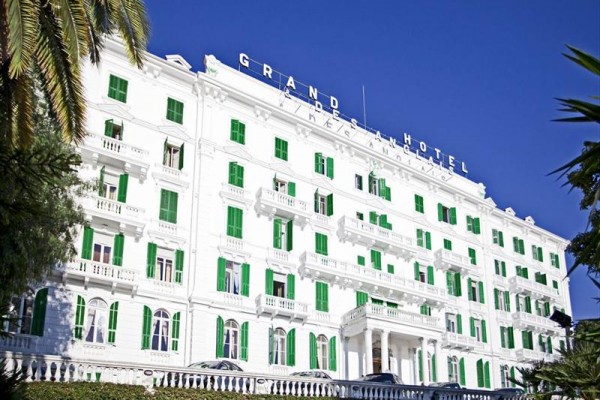 Historická budova bílého Grand Hotelu s výraznými zelenými okenicemi pod modrou oblohou.
