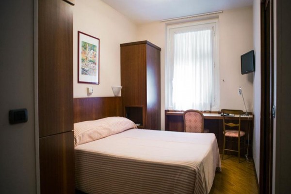 Jednoduše zařízený hotelový pokoj s postelí, stolkem, židlí a televizí na stěně.