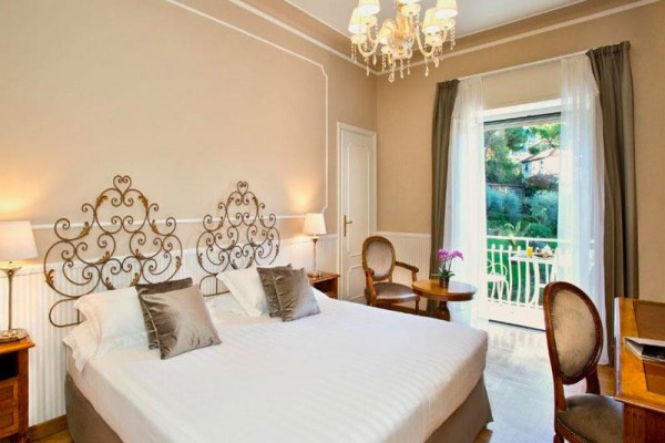 Luxusní hotelový pokoj s manželskou postelí, elegantním lustrem a balkonem s výhledem do zahrady.  