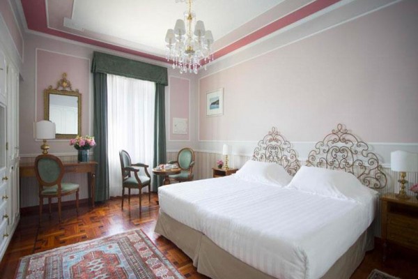 Elegantně zařízený hotelový pokoj s dvojlůžkem, křesly a stolem, v pastelových barvách.