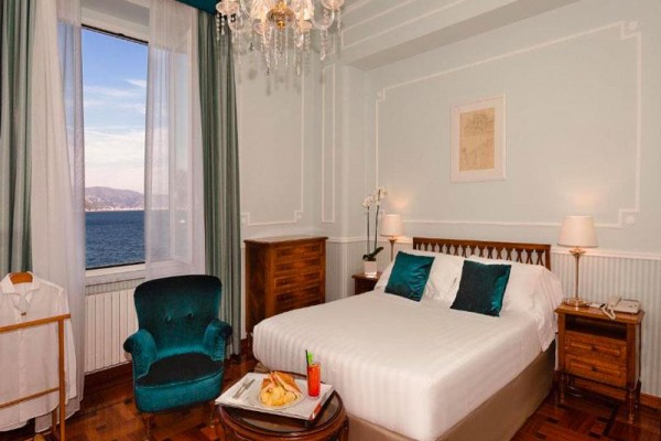Elegantní hotelový pokoj s výhledem na moře, vybavený lůžkem, křeslem a dřevěným nábytkem.  
