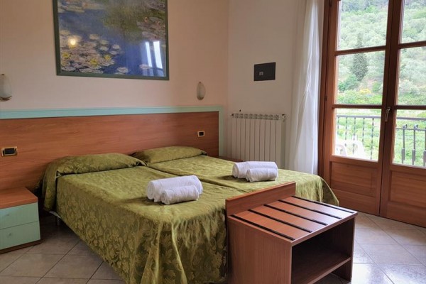 Pokoj se dvěma postelemi s přehozy, dřevěný nábytek a balkon s výhledem do zeleně.  