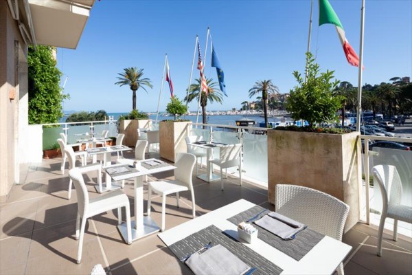 Terasa restaurace s bílými židlemi a stoly, výhled na moře a palmy, v pozadí vlajky.