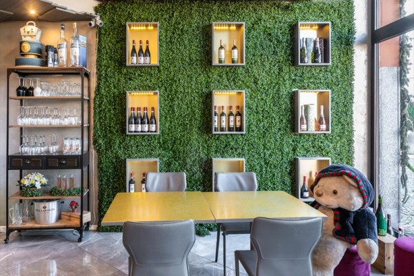 Restaurace s umělou zelenou stěnou ozdobenou lahvemi vína a stolem s plyšovým medvědem.