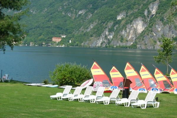 Bílé plastové lehátka na trávě v popředí, za nimi jezero s barevnými plachtami windsurfingů a zalesněné hory.