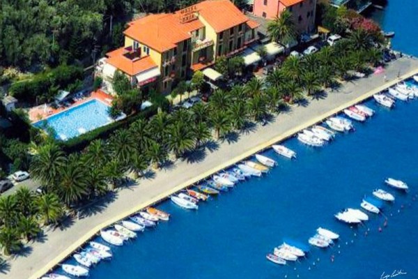 Hotel s bazénem poblíž pobřežní promenády lemované palmami a kotvícími loděmi.