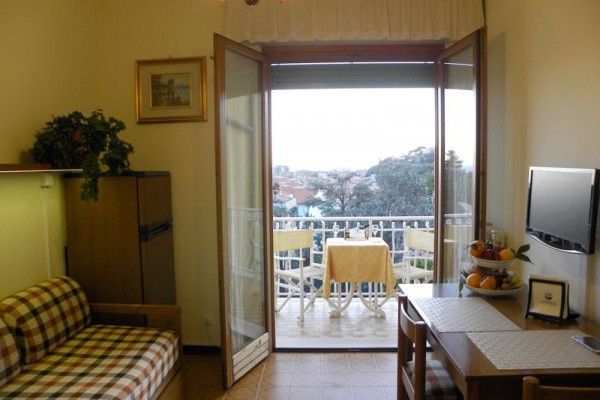 Otevřené dveře vedou na balkon s malým stolkem a židlemi, výhled na město v pozadí.  