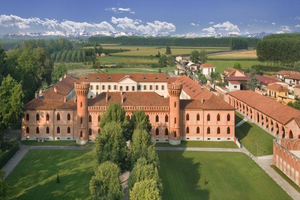 Historická cihlová budova s věžemi obklopená stromy a travnatými plochami na venkově.