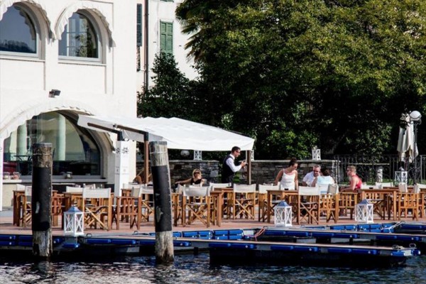 Venkovní restaurace u vody s lidmi sedícími u stolů a číšníkem obsluhujícím hosty.
