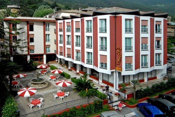 Čtyřpatrový hotel s venkovním posezením a červenobílými slunečníky.