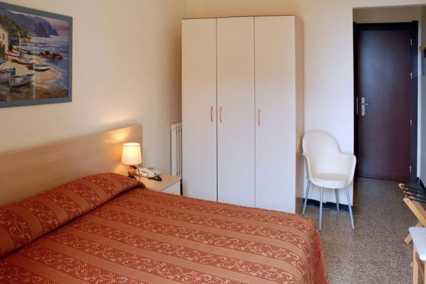 Hotelový pokoj s manželskou postelí, skříní, židlí a nočním stolkem s lampou.