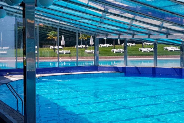 Krytý bazén s průhlednou střechou a venkovním výhledem na lehátka a slunečníky.