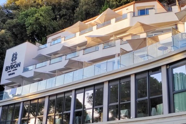 Moderní hotel se skleněnými balkony a slunečníky zasazený v zeleni.