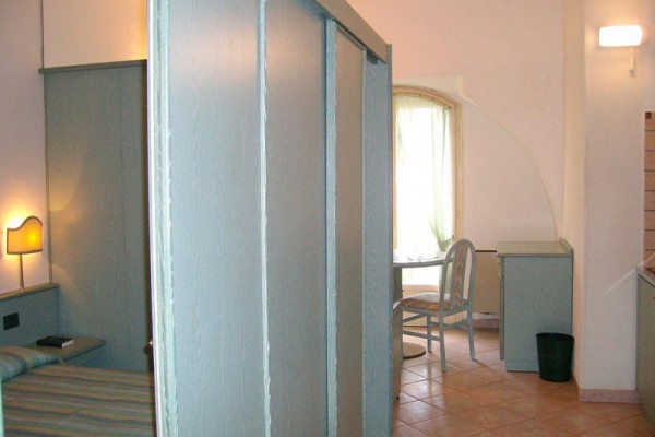 Jednoduše zařízený hotelový pokoj s postelí, lampou a menším stolem u okna.
