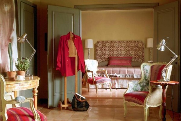 Elegantní ložnice se stylovým nábytkem a červenými doplňky, s otevřenými dveřmi do sousední místnosti s postelí.