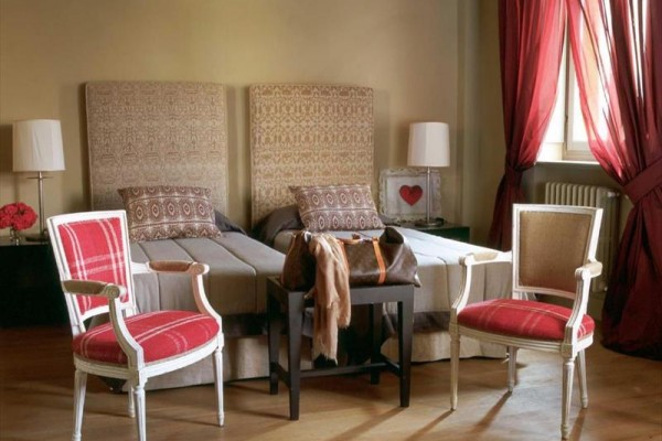 Elegantní ložnice s dvěma lůžky, dvěma červenými křesly a červenými závěsy u okna.
