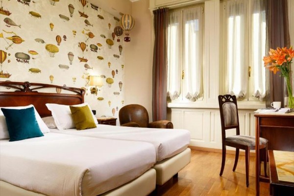 Pokoj v hotelu s dvěma jednolůžky, tapetou s horkovzdušnými balóny a okny s dlouhými závěsy.